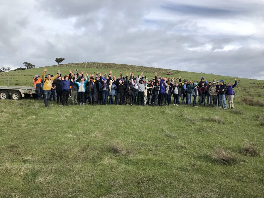 A crowd of planters waving from a green hillside at the Glenaroua planting weekend, a trailer of supplies beside them.