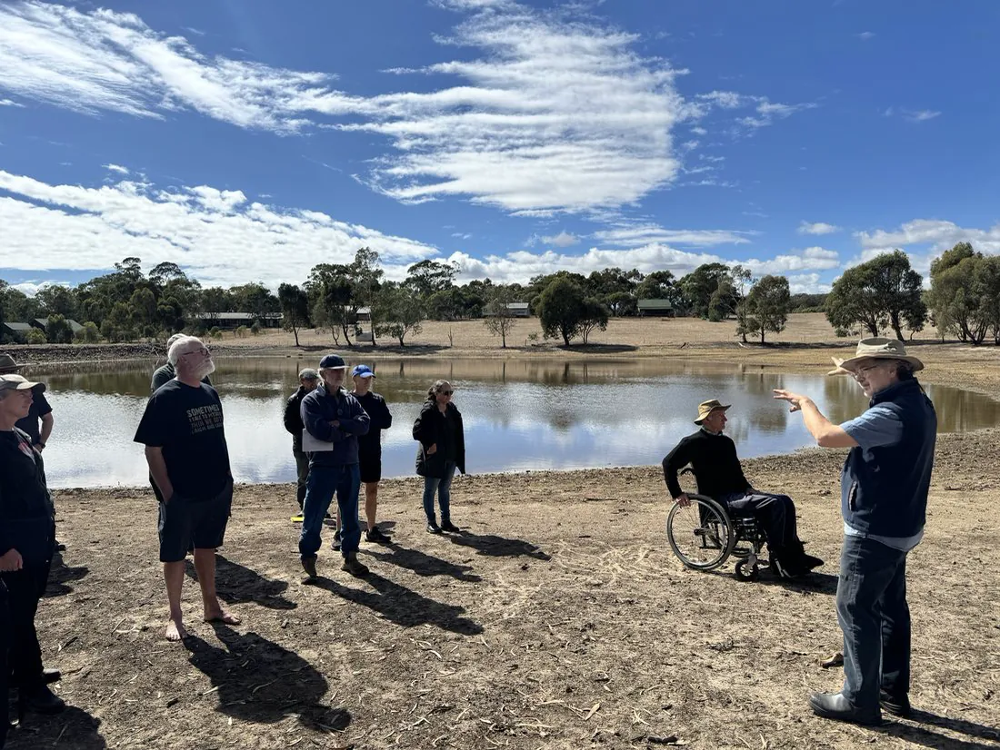 A group of landholders, one in a wheelchair, standing on the bank of the Lyndale Park dam while a man in a hat talks and points across the water.