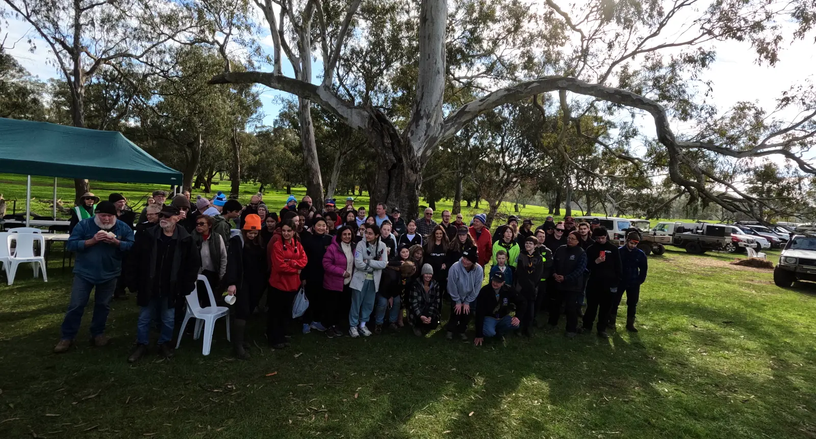 The volunteers of the June 2026 Glenaroua planting gathered for a photograph under a big old gum, a marquee and parked cars behind them.